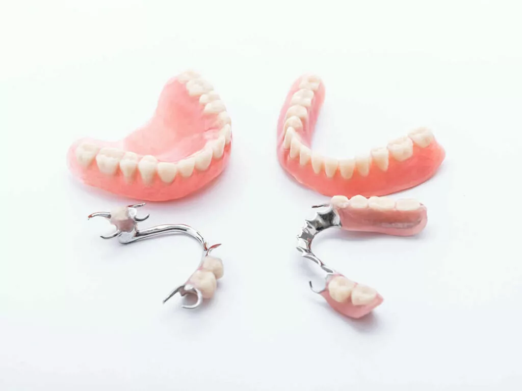Two full sets of dentures and two partial dentures placed on a white background. The full dentures are pink with white artificial teeth, and the partial dentures have metal clasps with a few artificial teeth on a pink base.