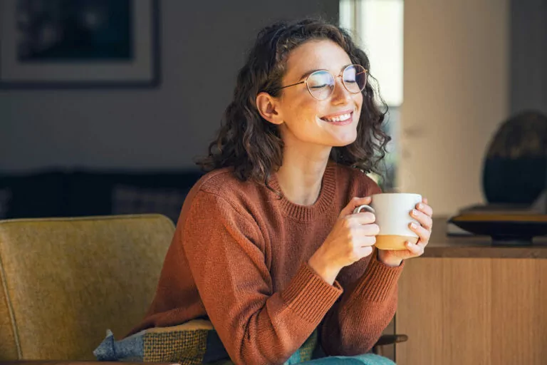The image shows a woman sitting on a chair, holding a white mug with both hands. The person is wearing a rust-colored sweater and has curly hair. The background includes a wooden table with a decorative object on it and a blurred interior setting, suggesting a cozy indoor environment.