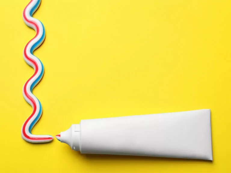 A white toothpaste tube on a bright yellow background with red, white, and blue toothpaste squeezed out in a wavy line.
