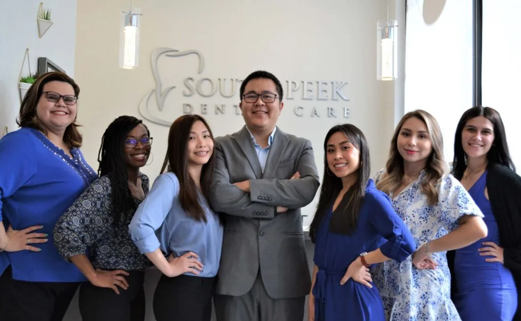 A group of seven people in professional attire standing in front of a 'SOUTHPEEK DENTAL CARE' sign. The central figure wears a gray suit with crossed arms, while the others pose with hands on their hips or by their sides. The background features two hanging lights and a window.