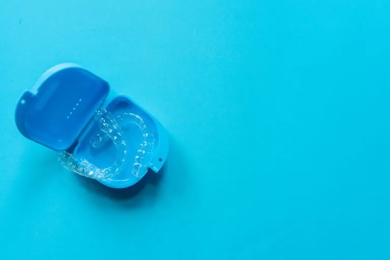 A blue plastic case containing clear dental aligners. The case is open, revealing the aligners inside. The background is a solid light blue color.