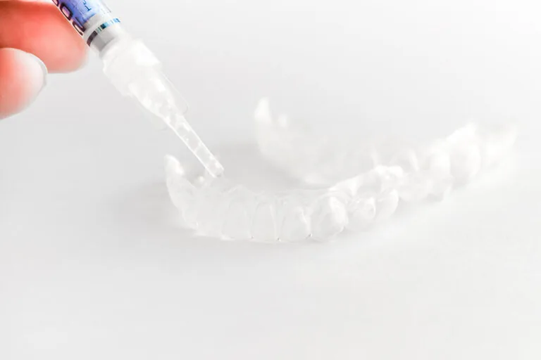 A syringe is being used to apply a substance to a clear dental aligner. Another clear dental aligner is visible in the background.