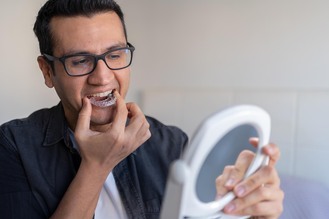 Person holding small mirror while adjusting clear dental aligner on teeth, focusing on proper placement in a clinical setting.